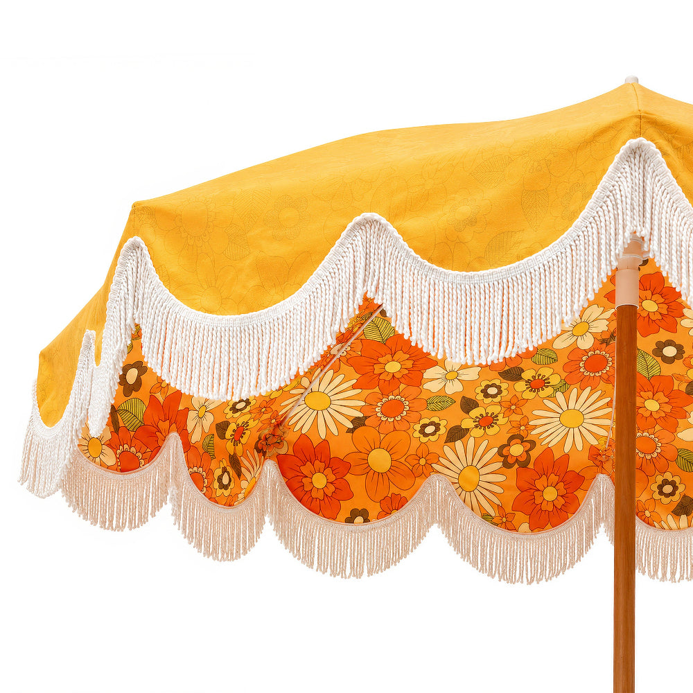 A close-up of a vintage-style beach umbrella canopy featuring a bright yellow top section, white fringe trim, and a retro orange floral scalloped lower section with daisy patterns, supported by a wooden pole.