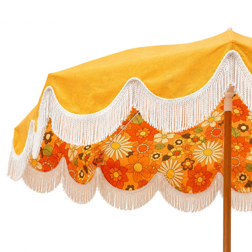 A close-up of a vintage-style beach umbrella canopy featuring a bright yellow top section, white fringe trim, and a retro orange floral scalloped lower section with daisy patterns, supported by a wooden pole.