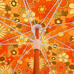 An overhead close-up of a floral umbrella canopy with a brown central pole. The fabric features a retro 1970s pattern of orange, yellow, and brown flowers with white ribs radiating outward.
