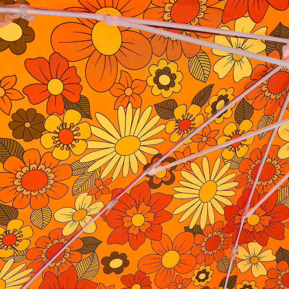 Close-up of a vibrant orange fabric with a retro 1970s floral pattern in yellow, red, and brown, overlaid with thin translucent pink cords.