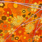 Close-up of a vibrant orange fabric with a retro 1970s floral pattern in yellow, red, and brown, overlaid with thin translucent pink cords.