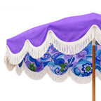 A close-up of a patio umbrella canopy showing a purple top section, white scalloped fringe trim, and a blue and purple floral patterned underside, with part of the wooden pole visible.