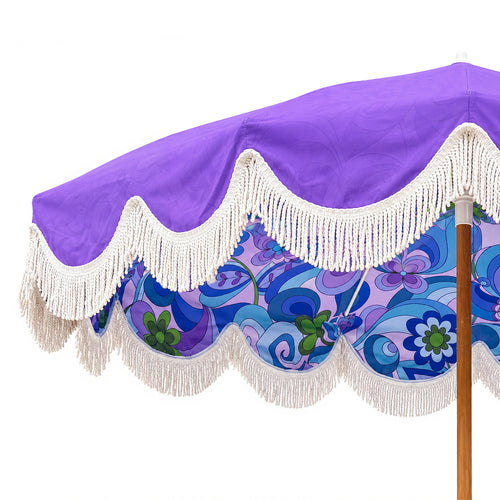 A close-up of a patio umbrella canopy showing a purple top section, white scalloped fringe trim, and a blue and purple floral patterned underside, with part of the wooden pole visible.