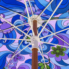 An overhead close-up of a vintage-style umbrella canopy with a retro 1960s psychedelic pattern in shades of blue, purple, and lavender, supported by white ribs and a brown wooden pole.