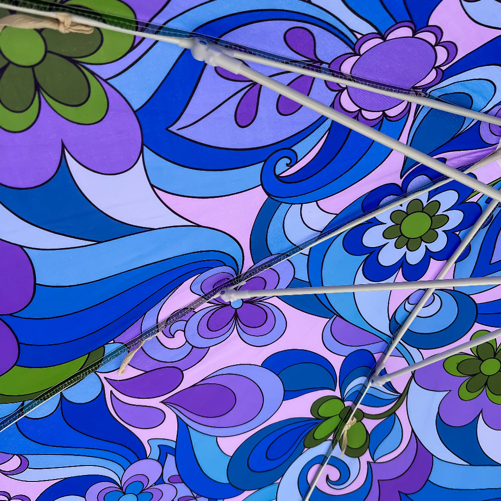 Close-up of a patterned umbrella canopy with a retro psychedelic design in shades of blue, purple, and green, with white metal ribs crossing diagonally.