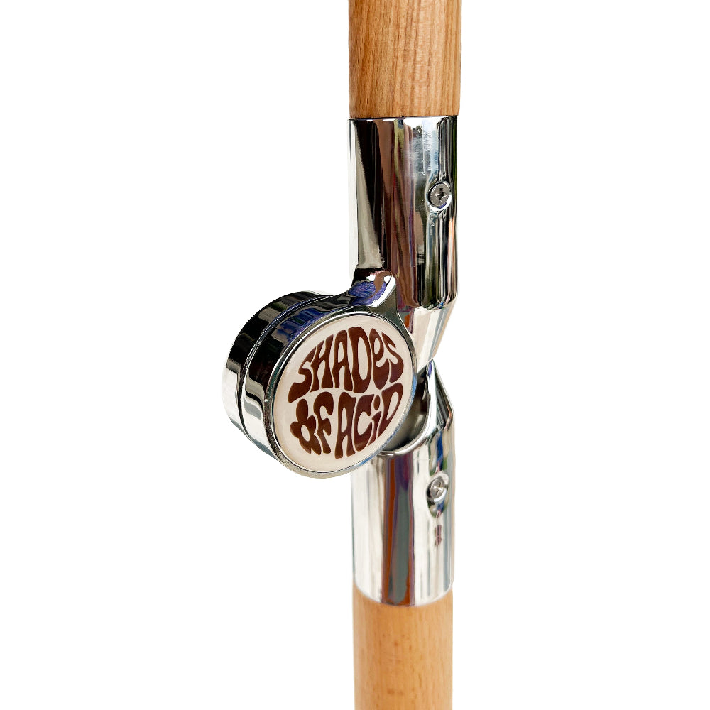 Close-up of a cane with a light wood shaft, polished chrome metal joints, and a circular white badge featuring the brand name 'Shades of Acid'.