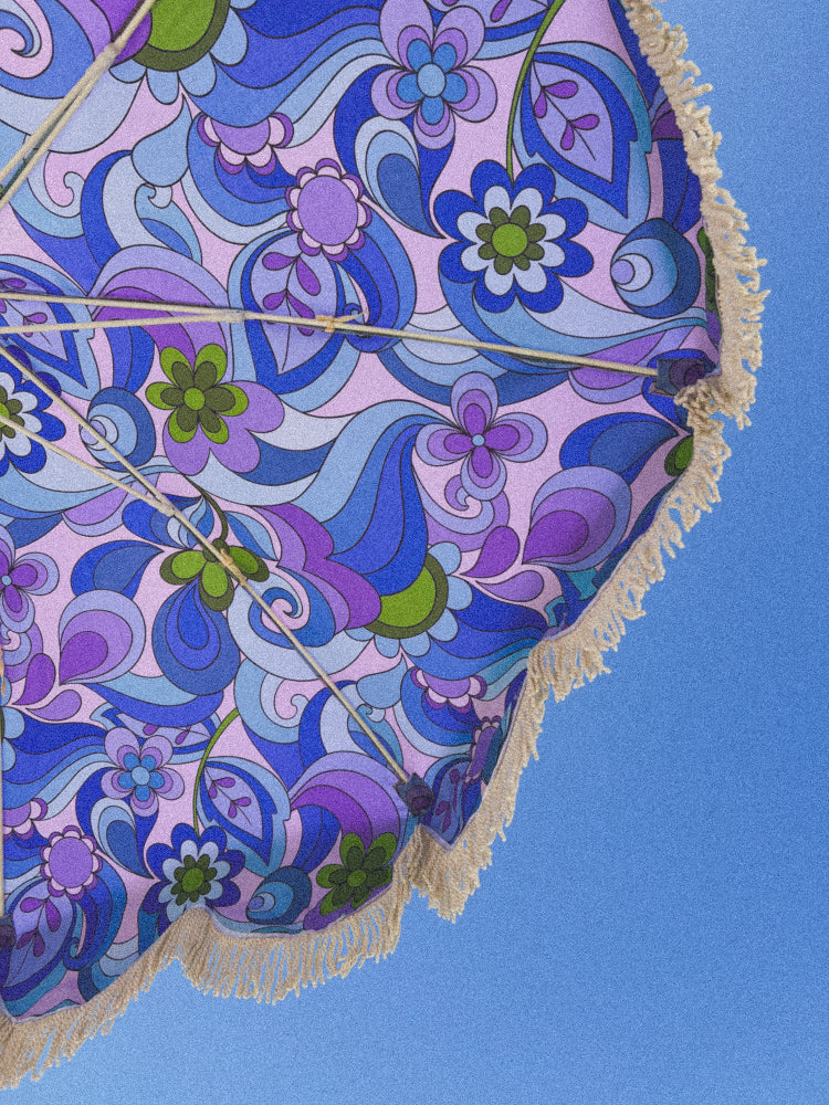 A close-up of a beach umbrella canopy with a retro 1970s floral and paisley pattern in shades of blue, purple, and pink, finished with a cream-colored fringe trim, set against a solid blue sky background.