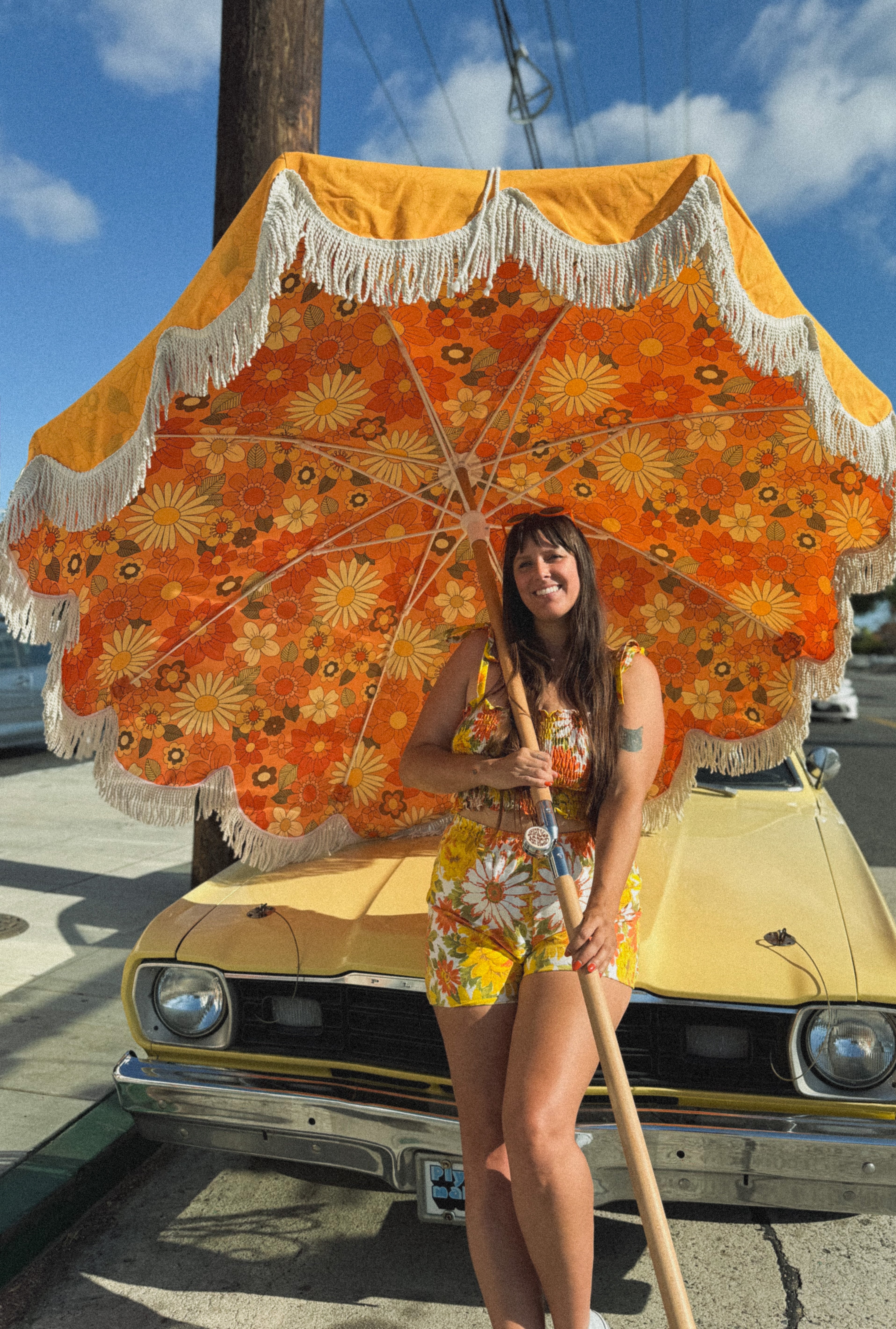 girl holding yellow fringe umbrella leaning against a classic car against a blue sky 